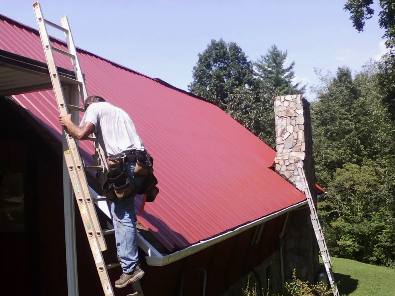 Roofer on a ladder beside a red metal roof and stone chimney — Soffit & Fascia Repair by Roofing Brothers in Sand Springs